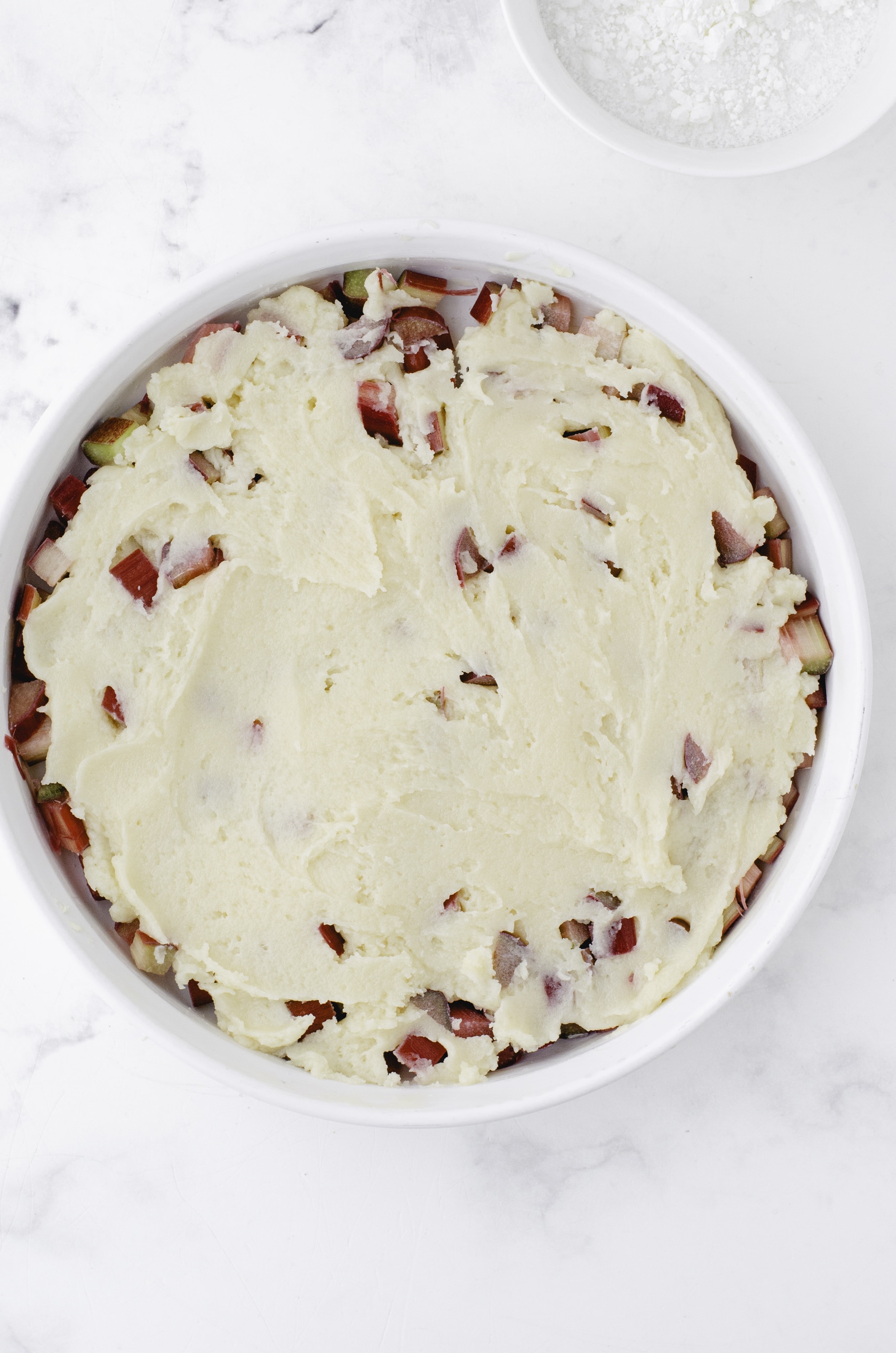 The cake batter smooth on top of diced rhubarb in a round white baking dish.