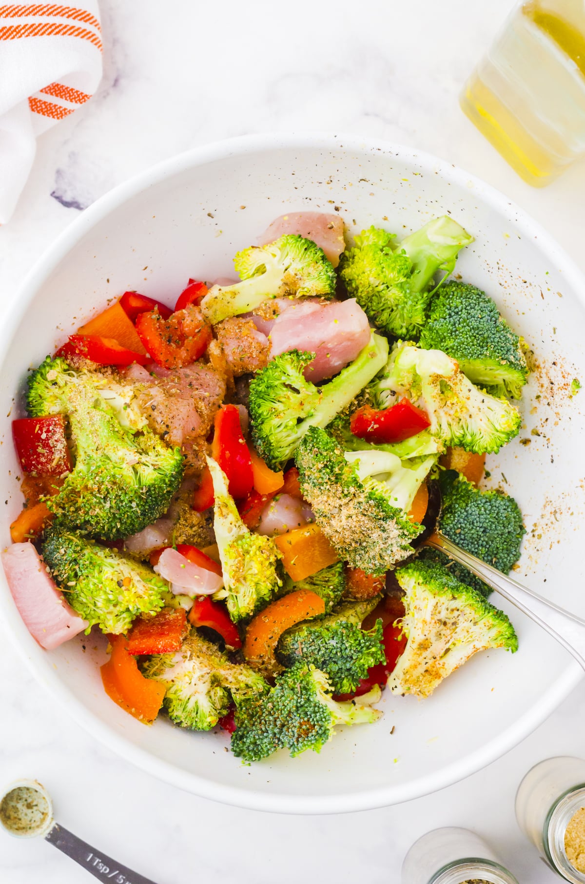 Broccoli, chicken, and peppers in a medium sized bowl being tossed with oil and spices for sheet pan dinner