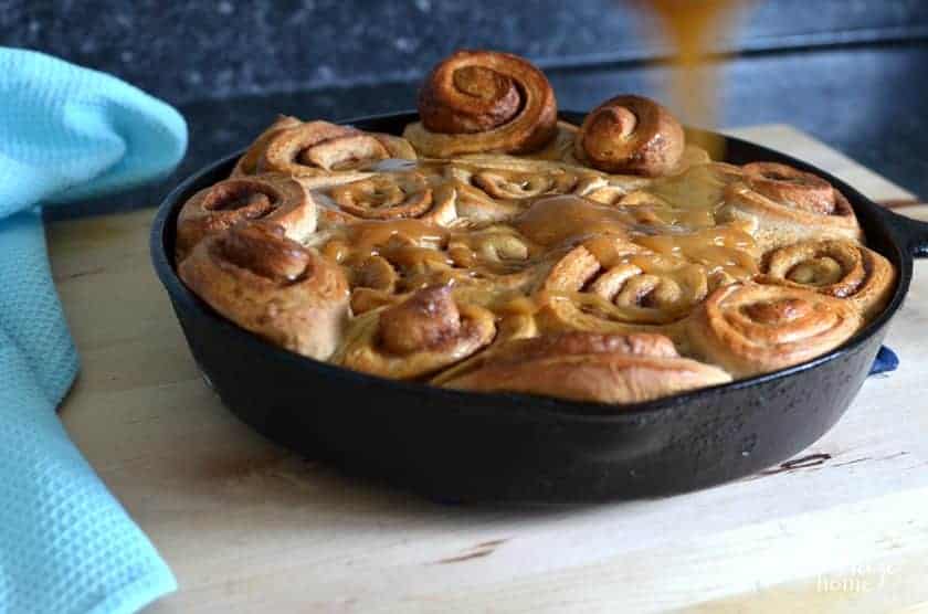 Caramel cinnamon roll caramel topping being poured on the top of a pan of skillet cinnamon rolls immediately after pulling them from the oven.