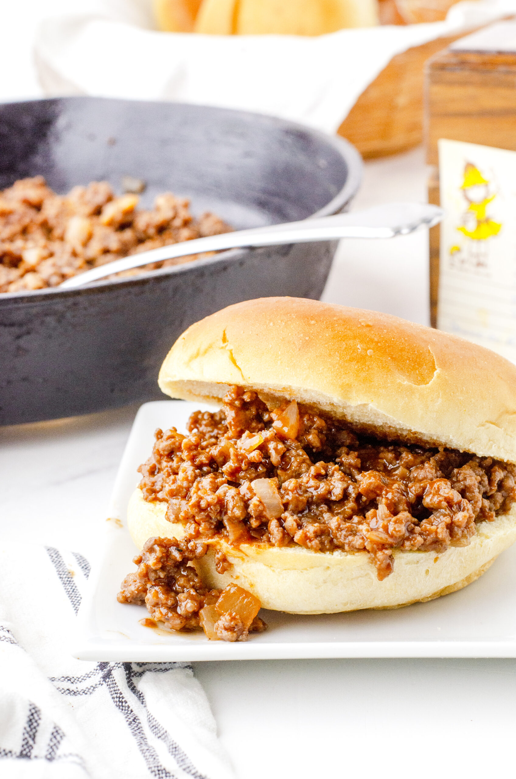 Old fashioned sloppy joes recipe served in a soft bun on a plate with a cast iron skillet in the background.