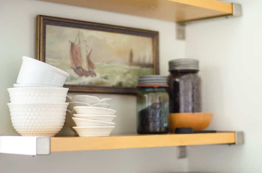 An open shelf with white bowls, antique jars filled with coffee beans, and an old art print of a ship on the wall behind it.