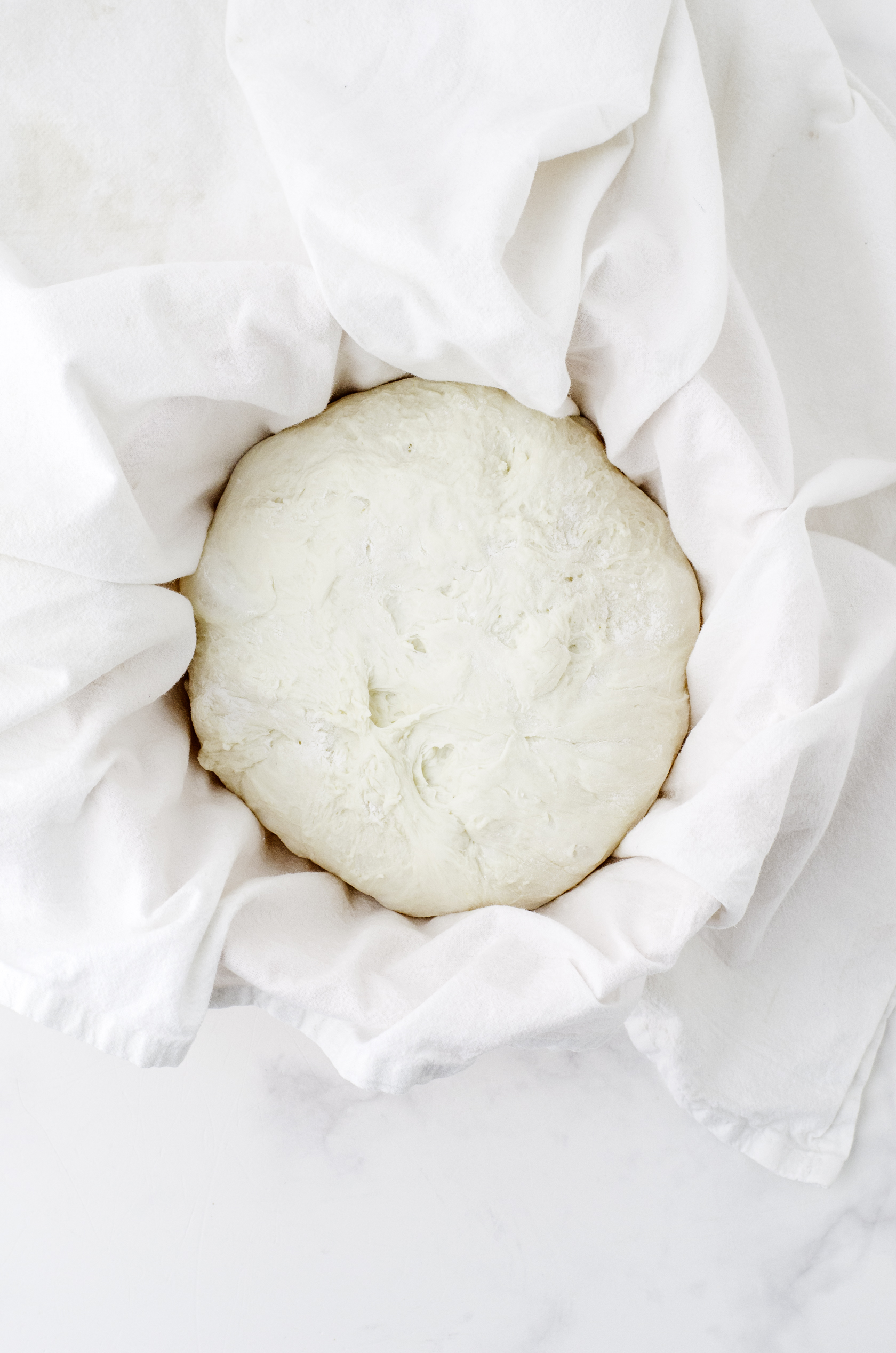 Sour dough bread formed and put in a bowl covered with a flour sprinkled dishtowel.
