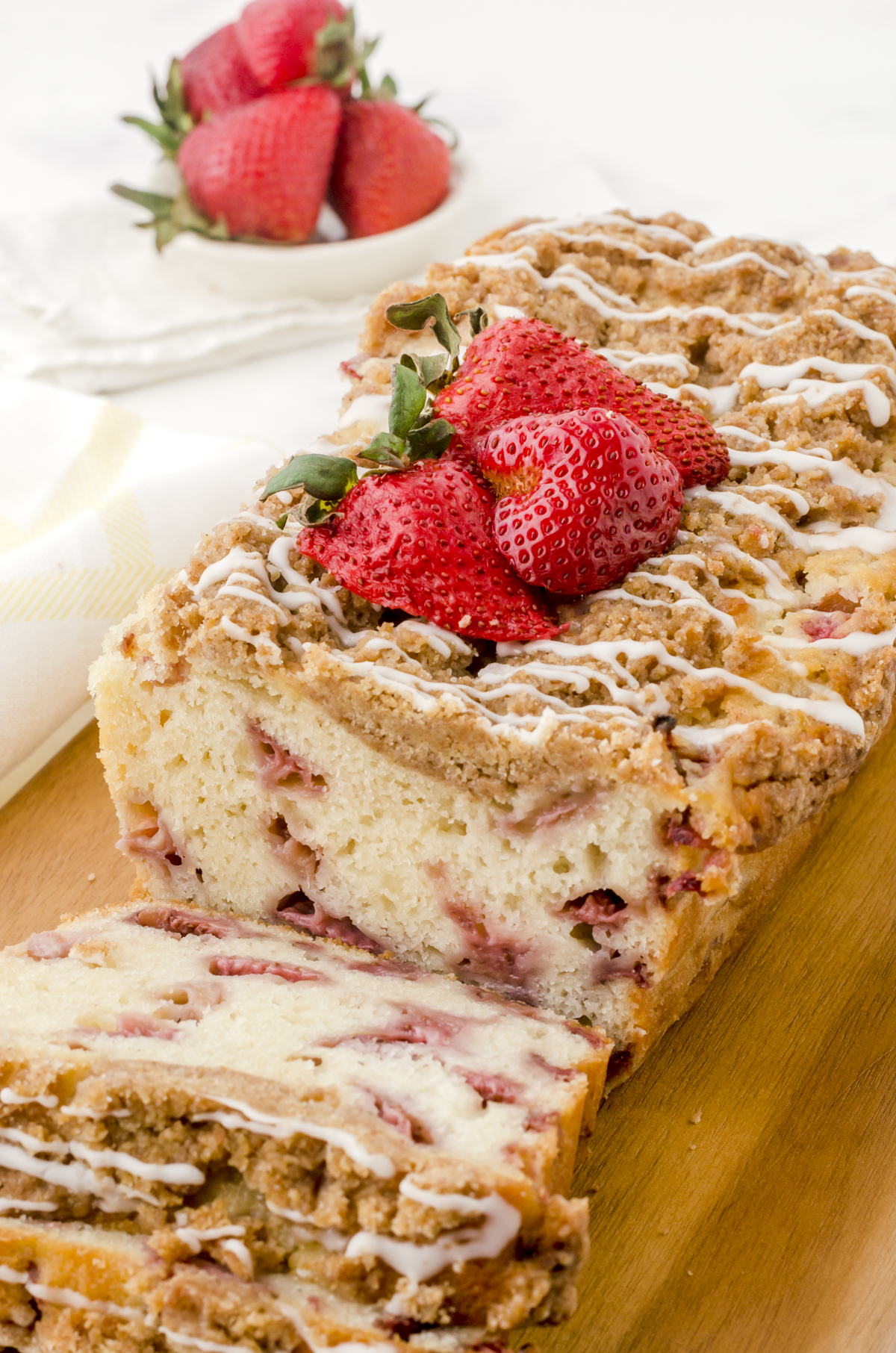 A loaf of strawberry bread with crumble topping and vanilla glaze on a wooden cutting board.