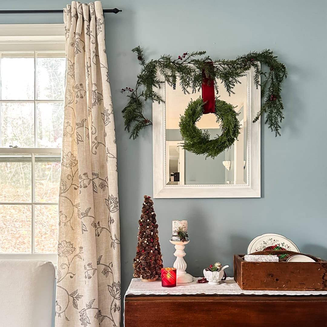 A cozy room with a window and floral curtains, a dresser topped with holiday decor, including a mini tree, candles, and dishes. A wall mirror is decorated with garland and a wreath hanging from a red ribbon.