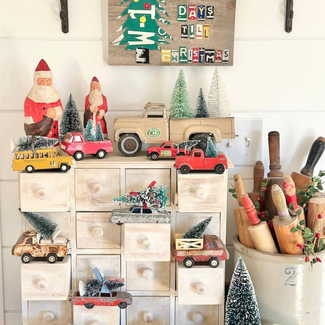 A festive display with vintage toy cars, trucks, and buses carrying mini Christmas trees, small Santa figurines, and bottle brush trees on a wooden cabinet. A holiday sign above reads “Days til Christmas.” Rolling pins are in a crock.