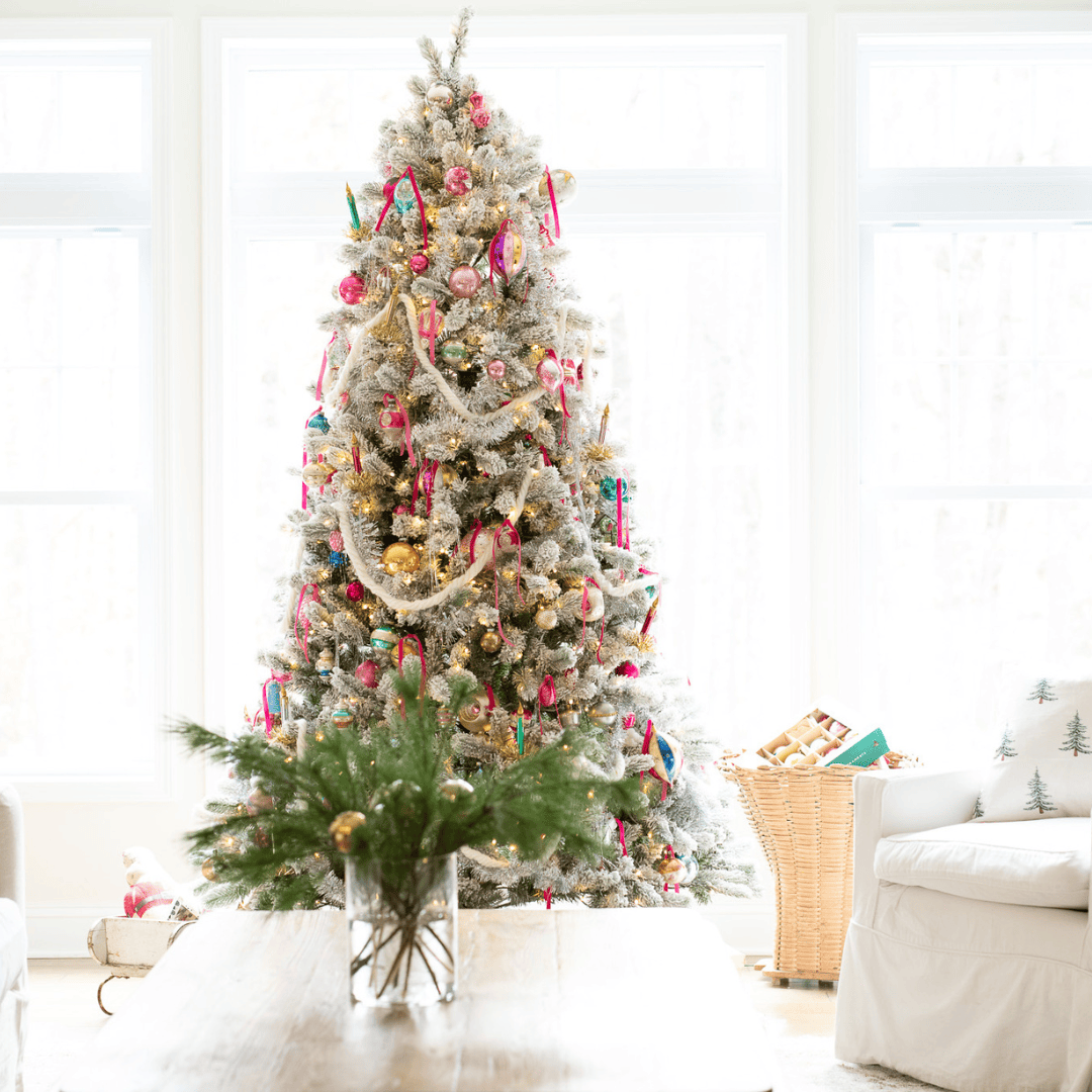 A brightly decorated Christmas tree with colorful ornaments and ribbons stands in a sunlit living room between two white armchairs. A vase with pine branches sits on a wooden coffee table in the foreground.