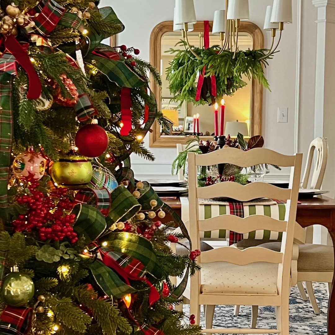 A decorated Christmas tree with red ornaments and plaid ribbons stands near a dining table set with candles, greenery, and plaid napkins. A greenery wreath hangs on a mirror, reflecting a festive, cozy holiday scene.