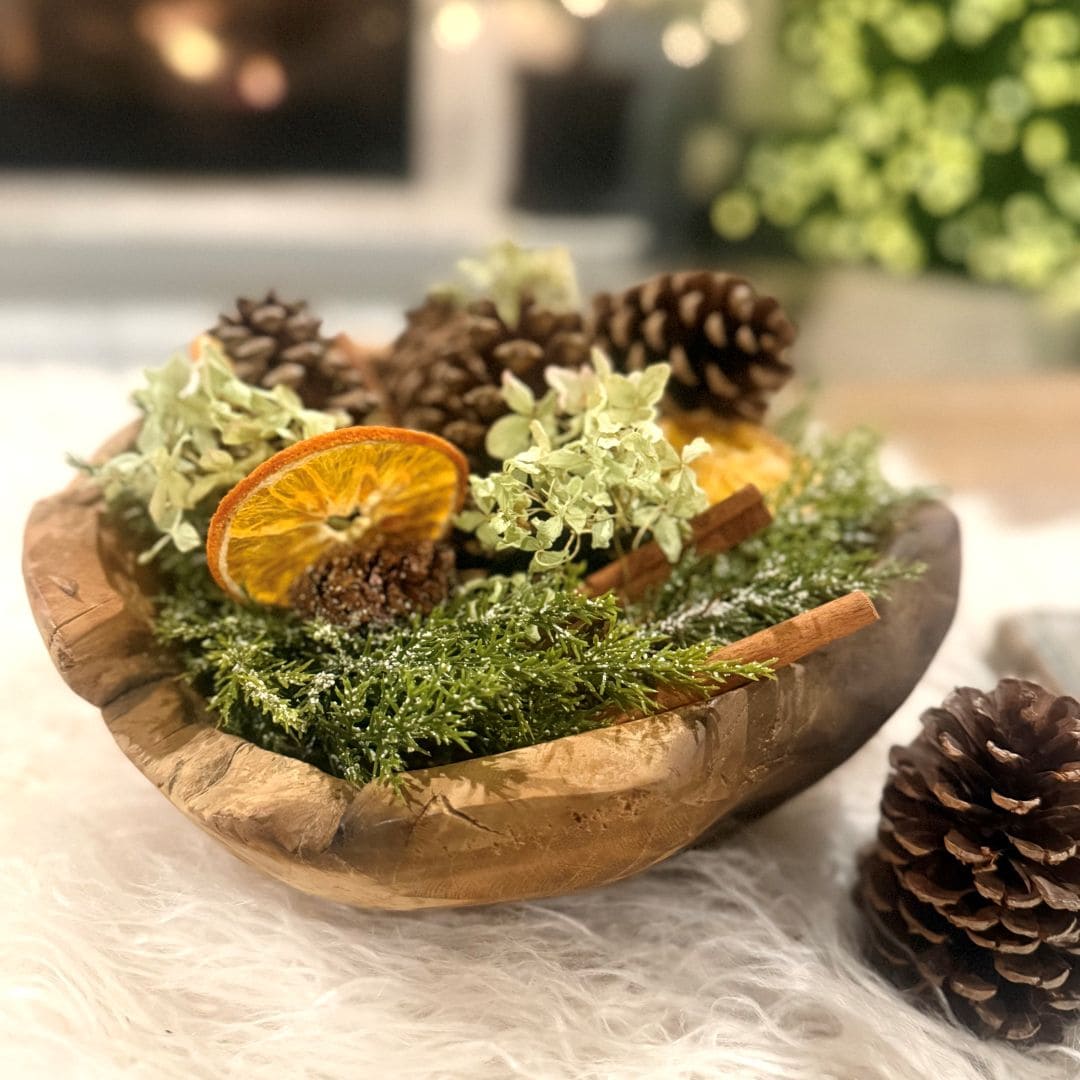 A wooden bowl filled with pinecones, dried orange slices, cinnamon sticks, green foliage, and decorative moss sits on a fluffy white surface, with pinecones scattered nearby.