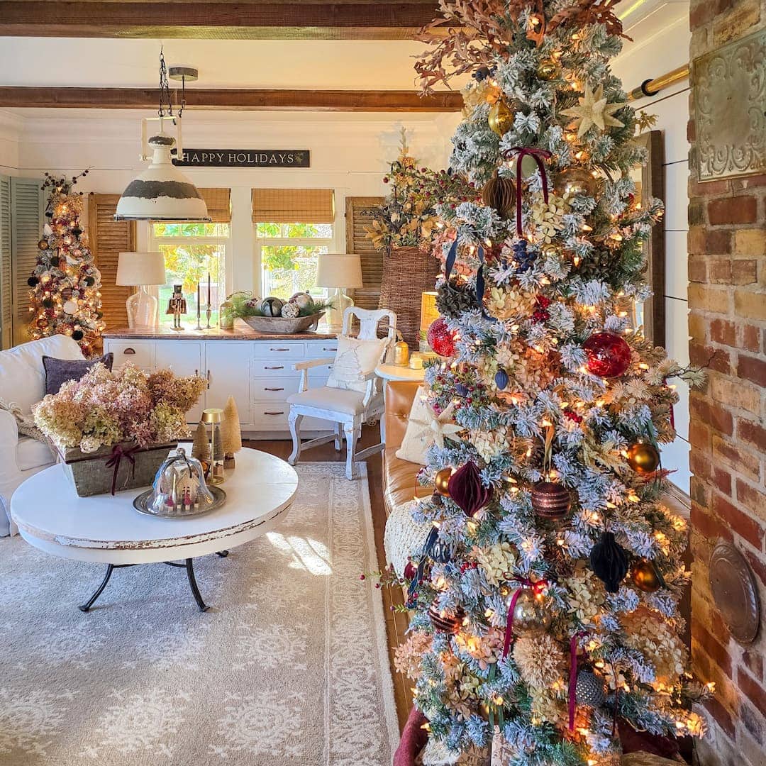 A cozy living room decorated for Christmas with a lit and ornamented Christmas tree, a festive wreath, a white sofa, a round coffee table with floral centerpieces, and sunlight streaming through the windows.