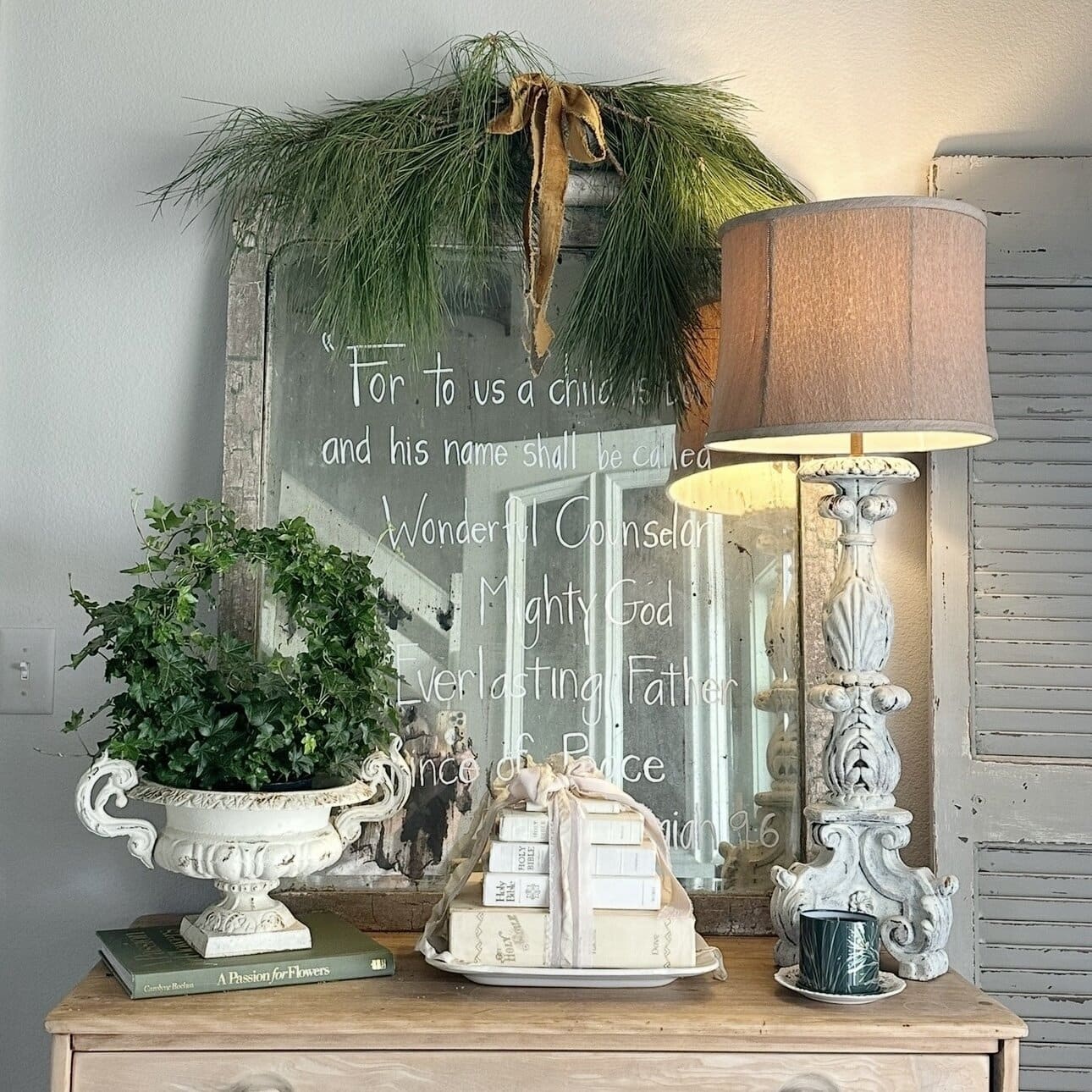 A decorated dresser with stacked books, a potted plant in an ornate planter, and a tall lamp. Behind, a mirror with a Christmas greenery wreath and a Bible verse is written in white script on the glass.