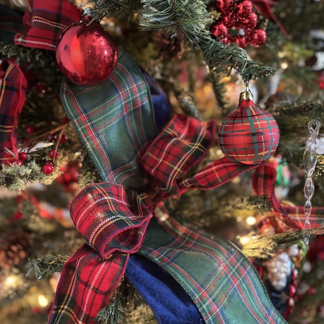 Close-up of a Christmas tree decorated with red and green plaid ribbon, red round ornaments, red berries, and warm white lights surrounded by green pine branches.