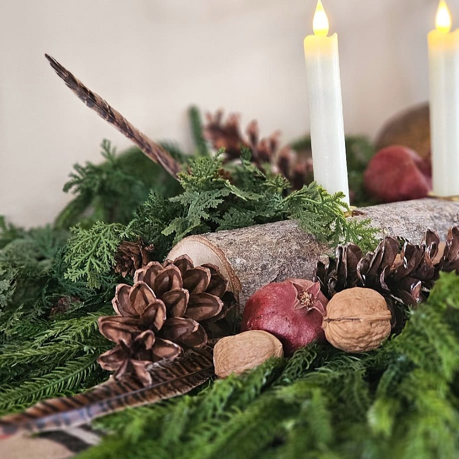 A festive arrangement with pinecones, walnuts, a pomegranate, green foliage, a wooden log, and two lit white candles, creating a natural holiday centerpiece.