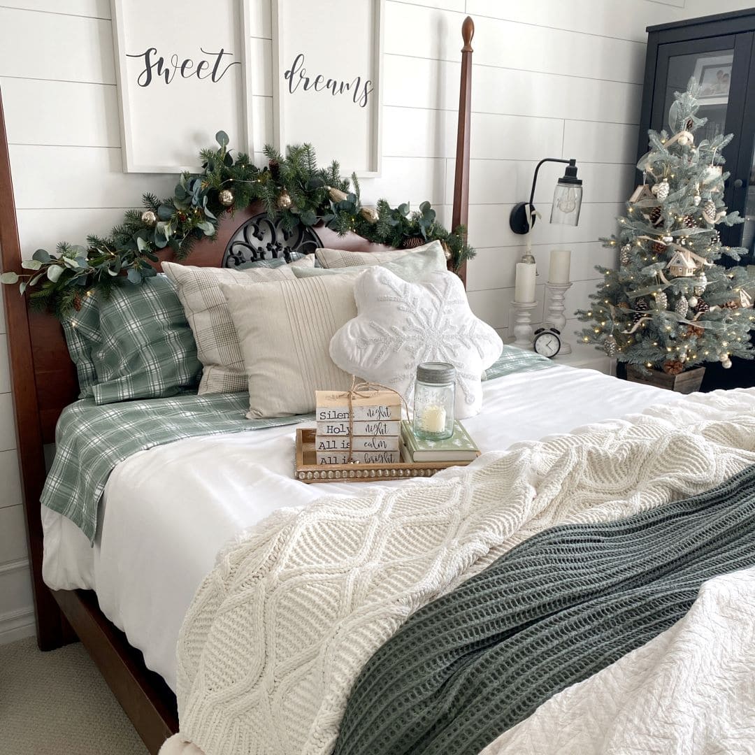 A cozy bedroom decorated for the holidays with green-and-white bedding, a knit blanket, Christmas garland on the headboard, a small decorated Christmas tree, and framed "sweet dreams" art above the bed.