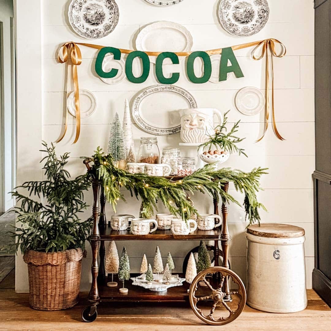 A festive cocoa bar cart decorated with greenery, vintage mugs, jars, ceramic trees, and a "COCOA" banner. Plates are hung on the wall, and a potted evergreen sits to the left of the cart.