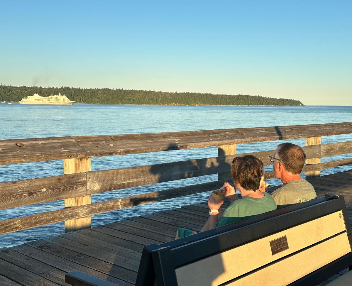 man and woman on a park bench eating icing cream with wooden spoons watching a cruise ship go by