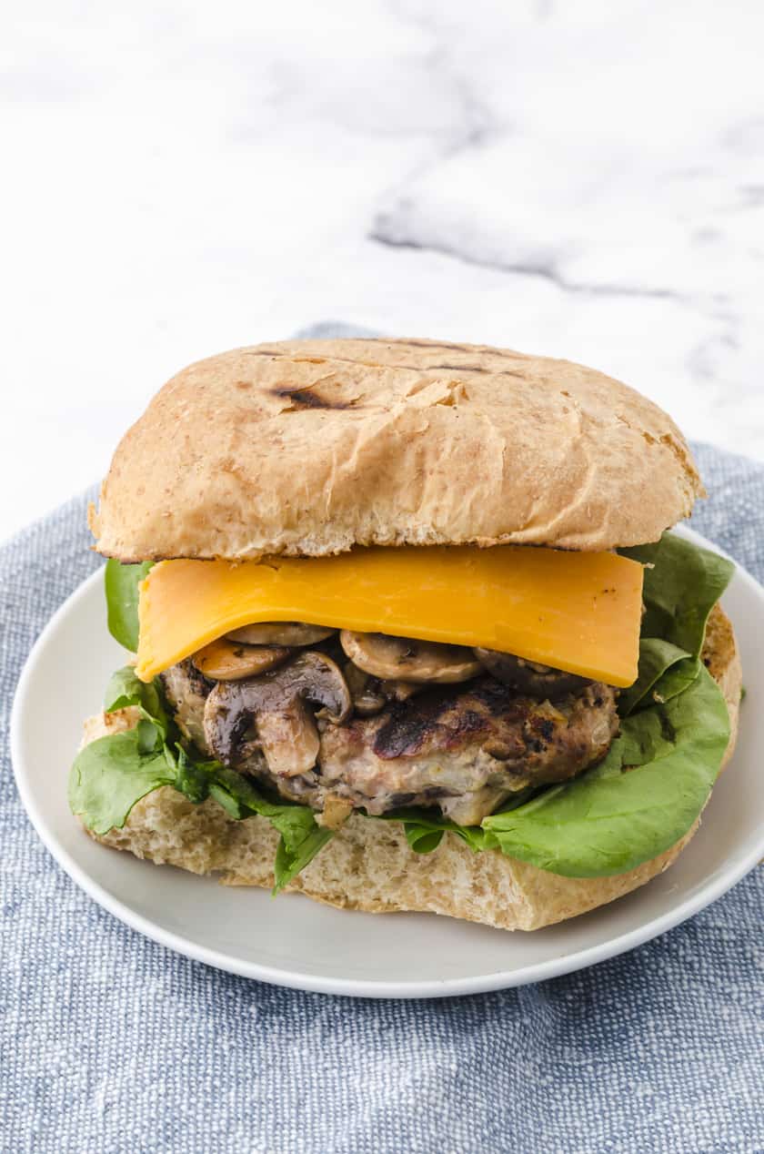 a grilled turkey burger on a plate with sauted mushrooms, lettuce, cheese and a toasted bun.