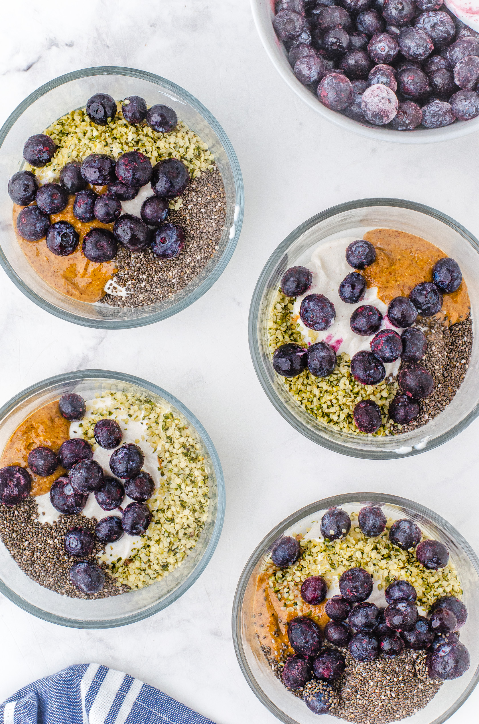 4 serving bowls with greek yogurt almond butter, chia seeds, hemp seeds and frozen blueberries in them.