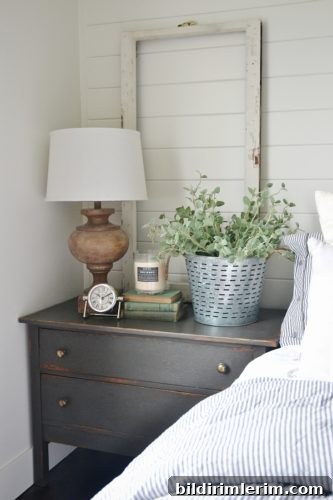 Stylish Modern Farmhouse Bedroom Ideas 6 A farmhouse style nightstand beside a bed with an old window frame propped up on it and a galvanized bucket of greenery, stack of books, candle and lamp on it.