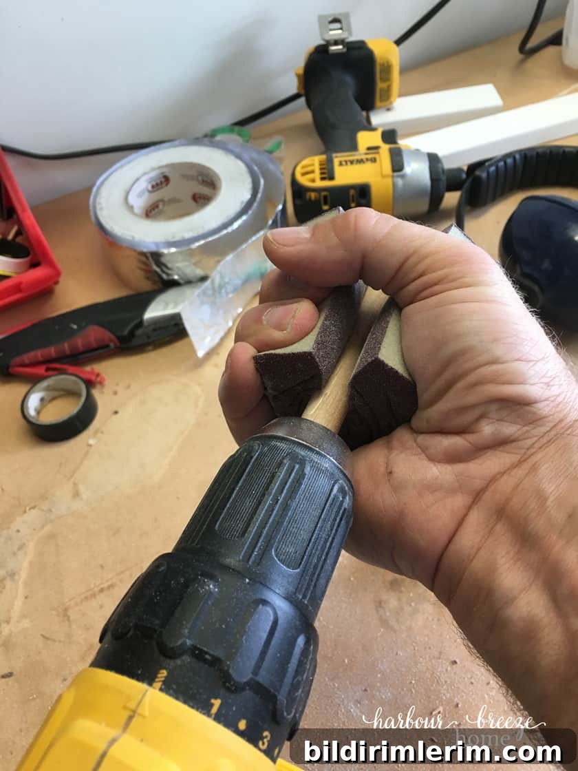 A dowel attached to the end of a drill with a sanding sponge squeezed around it with a man's hand.