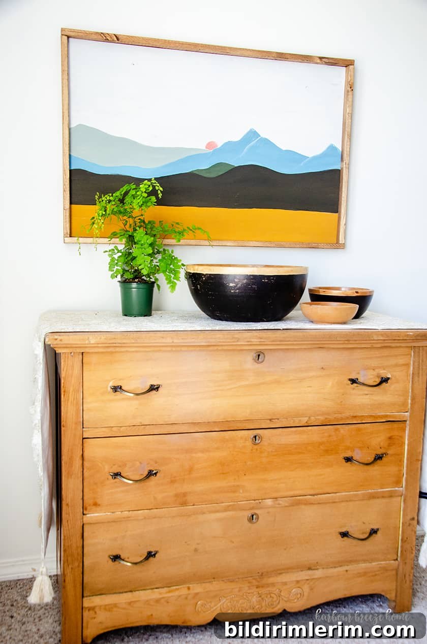 An easy mountina picture in a boho style hung above a wooden dresser with plants and wooden bowls in front of it.