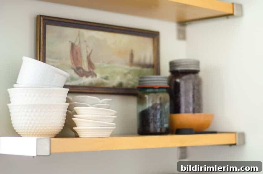 Budget-Friendly Small Kitchen Remodel Ideas 9 An open shelf with white bowls, antique jars filled with coffee beans, and an old art print of a ship on the wall behind it.