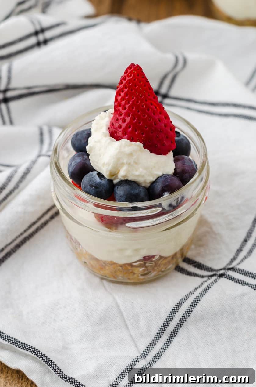 No bake cheesecake in mini mason jar with a strawberry on the top surrounded by blueberries. It's a red white and blue dessert!