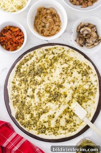 pesto sauce being spread over pizza dough on a stoneware pan