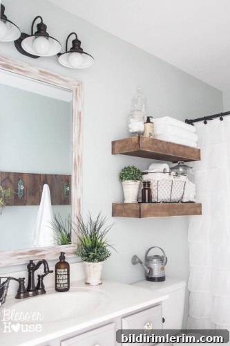 A bathroom design with farmhouse style similar to joanna gaines bathroom. It has 2 wooden shelves above the toilet, farmhouse style lighting about the vanity, and accessories like plants, wire baskets, and glass jars filled with cotton balls.