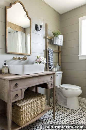 A bathroom design in the farmhouse style similar to what you would see on a Joanna Gaines Fixer Upper show. It has shiplap on the walls, a wooden framed mirror, and a ladder propped behind the toilet with a basket filled with towels attached.