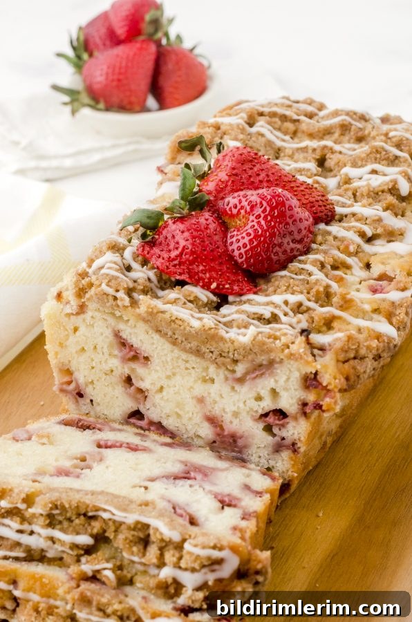 A loaf of strawberry bread with crumble topping and vanilla glaze on a wooden cutting board.