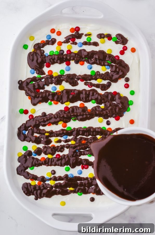 How to Make Oreo Ice Cream Cake with Oreo Crust 7 hot fudge sauce being drizzled over the m&ms layer of an oreo ice cream cake