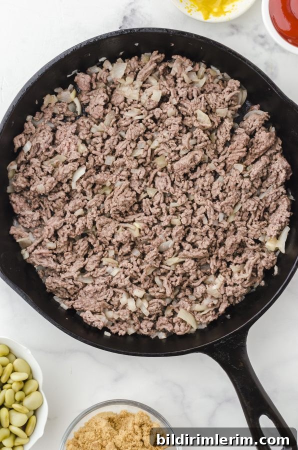 Browned ground beef  and onions in a cast iron skillet ready to be put in a crock pot for this baked beans recipe.