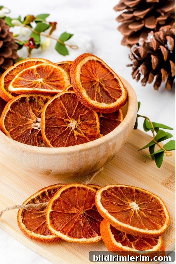 Dried orange slices in a wooden bowl sitting on a wood board surrounded by pine cones and faux greenery as an illustration of dried orange slices decor