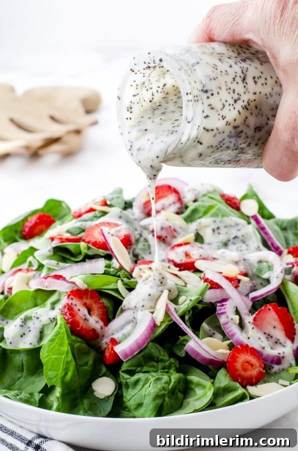 Creamy poppy seed dressing being drizzled onto a spinach and strawberry salad.