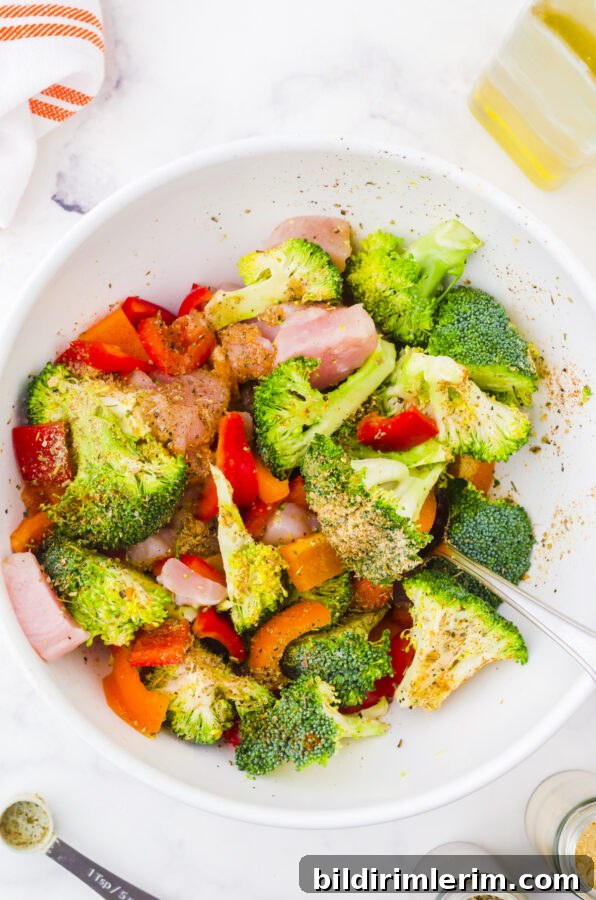 Broccoli, chicken, and peppers in a medium sized bowl being tossed with oil and spices for sheet pan dinner