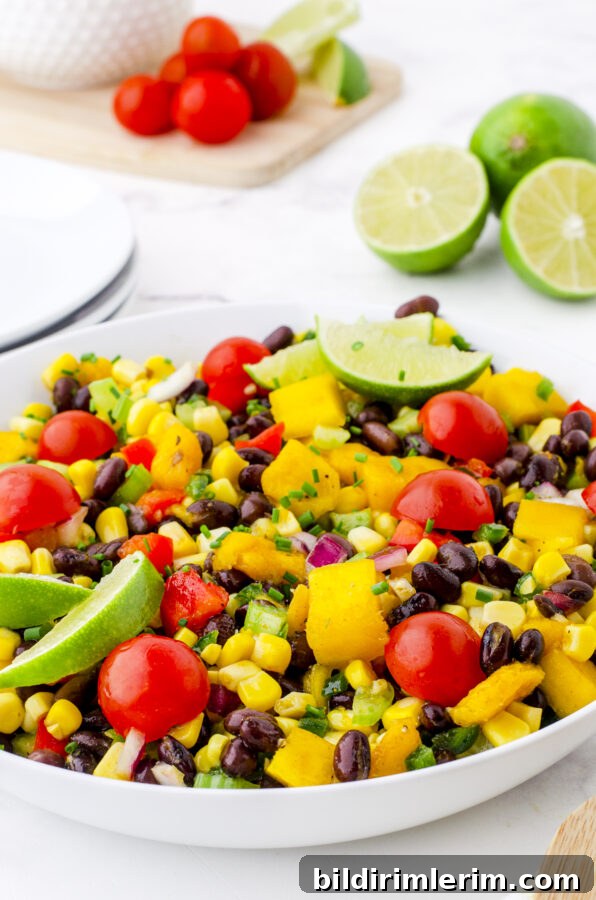 A bowl of mango salad with black beans garnished with lime slices, cherry tomatoes, and fresh chives.