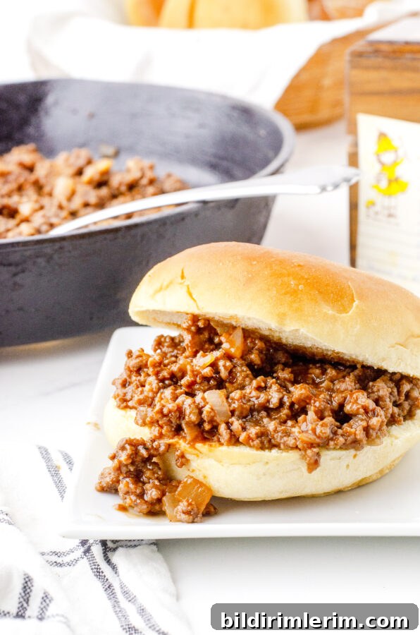 Old fashioned sloppy joes recipe served in a soft bun on a plate with a cast iron skillet in the background.