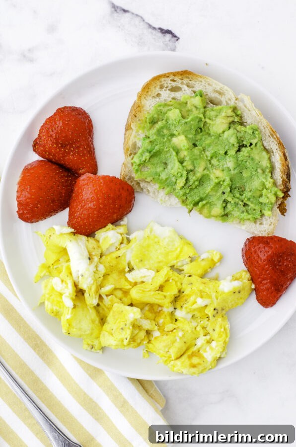 Mashed avocados on a piece of sourdough toast on a plate with scrambled eggs and strawberries.