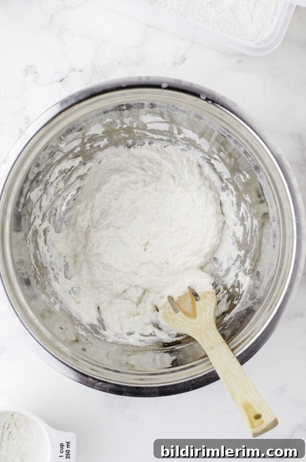 Flour and water being stirred into sourdough starter with a wooden spoon with holes in it. Step 4 of feeding sourdough starter.