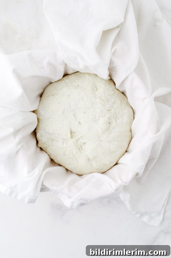 Sour dough bread formed and put in a bowl covered with a flour sprinkled dishtowel.